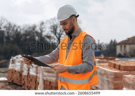 Holding notepad with documents. Construction worker is at job with bricks outdoors.