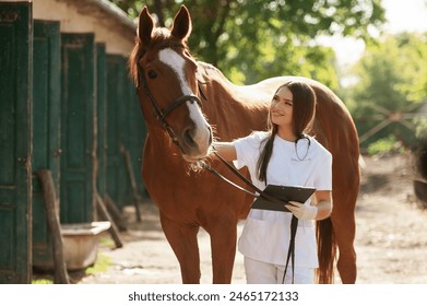 Holding notepad. Female vet is with horse outdoors at the farm at daytime. - Powered by Shutterstock