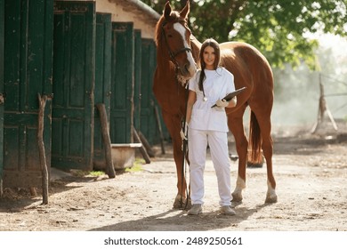 Holding notepad. Female vet is with horse outdoors at the farm at daytime. - Powered by Shutterstock