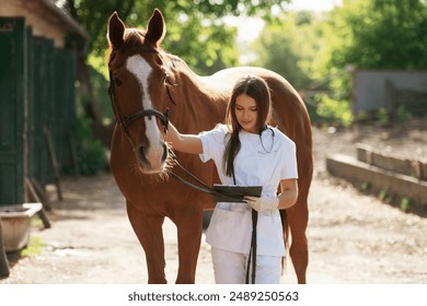 Holding notepad. Female vet is with horse outdoors at the farm at daytime. - Powered by Shutterstock