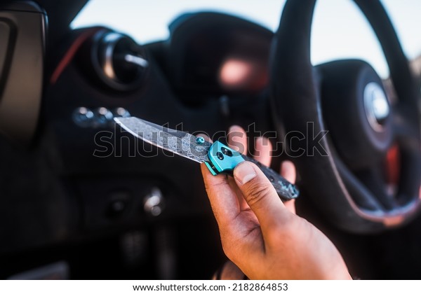 Holding a open pocket knife in hand in front of a\
steering wheel of a car