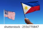 Holding Philippine flag waving under cloudy sky. Fly in the sky. Intentionally blurred photo of American flag in the distance