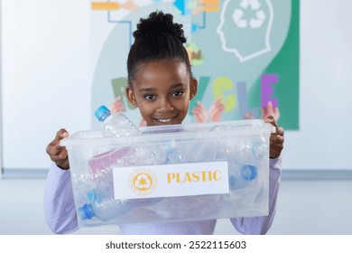 Holding plastic recycling bin, african american girl smiling in school classroom promoting recycling. sustainability, environment, eco-friendly, waste management, green - Powered by Shutterstock