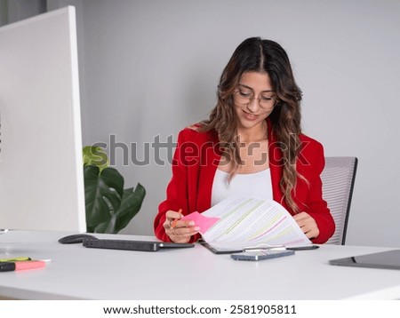 Holding reading documents, young smiling confident business woman sit at office desk holding reading documents. Concentrated 20s entrepreneur female worker employee wear red formal jacket.