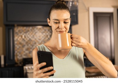 Holding smartphone and drinking coffee, woman enjoying morning in kitchen. Morning routine, coffee break, relaxation, home life, daily routine, technology - Powered by Shutterstock