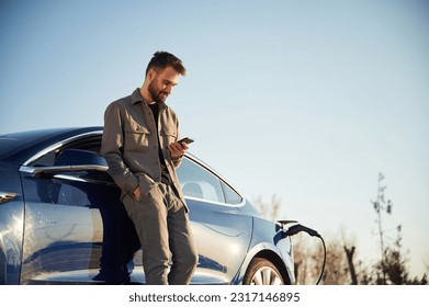 Holding smartphone in hand. Man is standing near his electric car outdoors. - Powered by Shutterstock