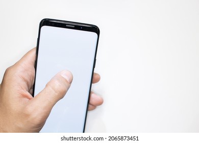 Holding Smartphone On White Background. Man's Hand Scrolling Smartphone With White Screen As Template. White Background And Empty Smartphone Frame. Thumb Finger On Telephone Screen