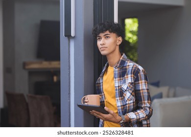 Holding tablet and coffee mug, teenage boy standing and looking outside, copy space. Technology, casual, contemplation, lifestyle, relaxation, indoors - Powered by Shutterstock