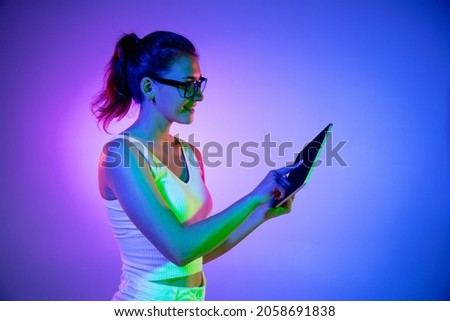 Holding tablet, crazy excited. Caucasian young woman's portrait on black studio background in neon light. Beautiful female model in white shirt. Concept of human emotions, facial expression, sales, ad