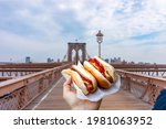 Holding two hot dogs in NYC on the Brooklyn Bridge .