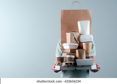 Holding various take-out food containers, pizza box, coffee cups in holder and paper bag, close-up. Light grey background, place to insert your text. Delivery man.