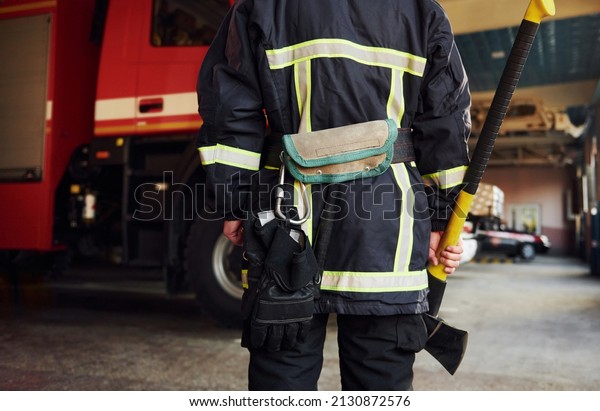 Holds axe in hands. Male firefighter in protective\
uniform standing near\
truck.