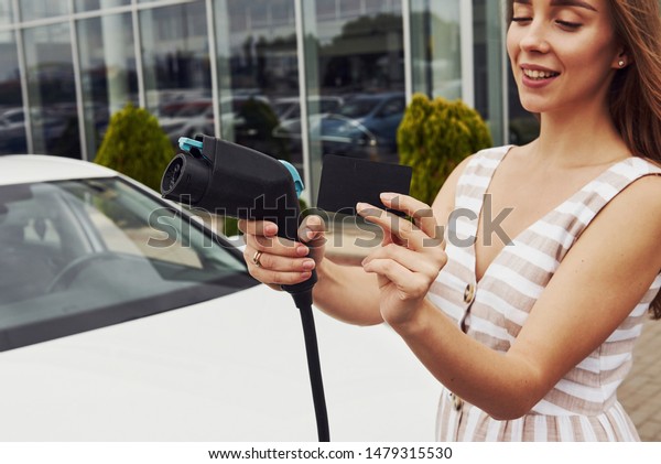 Holds black discount card.\
Woman on the electric cars charge station at daytime. Brand new\
vehicle.