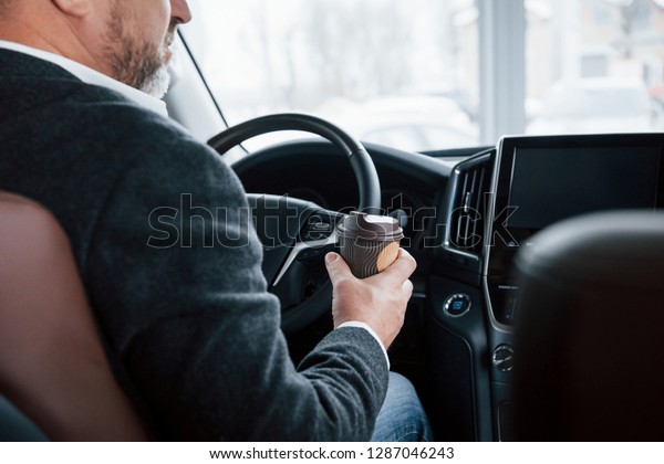 Holds cup with coffee. View\
from behind of senior businessman in official clothes inside of\
modern car.