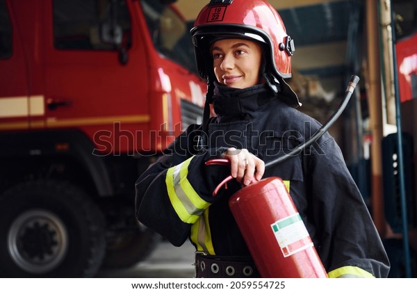 Holds extinguisher in hands. Female\
firefighter in protective uniform standing near\
truck.