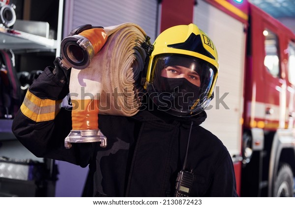 Holds hose. Male firefighter in protective uniform\
standing near truck.