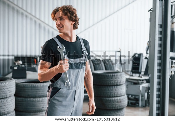 Holds wrench in hand. Adult man in grey\
colored uniform works in the automobile\
salon.