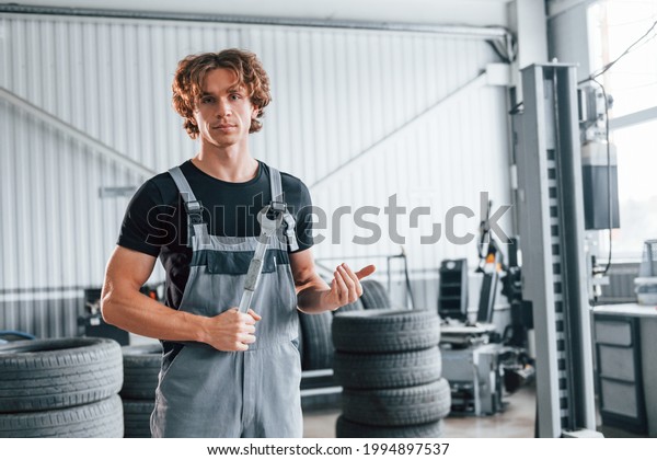 Holds wrench in hand. Adult man in grey\
colored uniform works in the automobile\
salon.