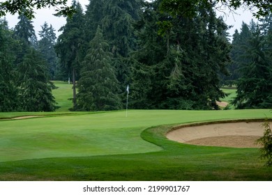 A Hole In A Golf Course Is Seen In Early Morning. No People Are Around. We Can See The Flag, Trees, And Part Of A Sand Trap.