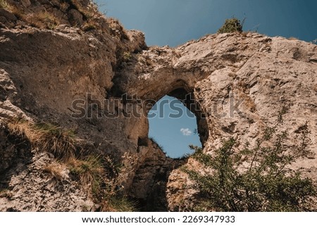 Hole in old rock with trees, grass, flowers and blue sky view. Natural attraction of Balkans. Dupni kamen is tourist landmark of Stara planina mountain range. Serbian natural site in Balkan Mountains.