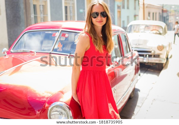 Holguin / Cuba\
10.10.2018: Beautiful woman in red dress is standing near the red\
car on the street of\
Cuba