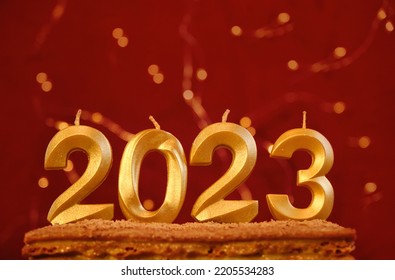 Holiday Background Happy New Year. Minimalistic Horizontal Red Background With Bright Yellow Bokeh Garland And Copy Space. Numbers 2023 Made By Gold Candles. Candles Without Fire Stuck In Cake.