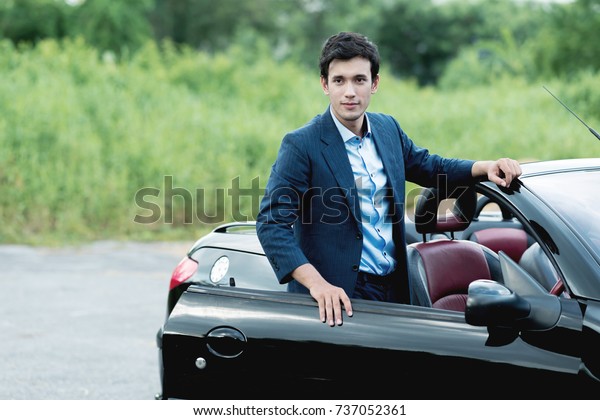 Holiday Car\
rent and road trip concept. Handsome white man standing on a side\
of black convertible with nature background, looking into camera,\
ready for a road trip and long\
drive.