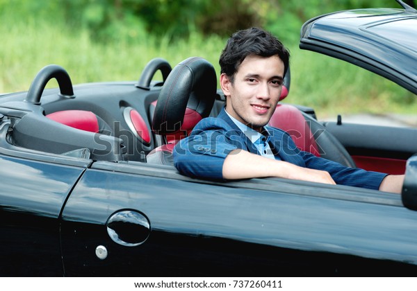 Holiday Car rent and road trip concept.\
Handsome white man inside a black convertible with nature\
background, looking into camera, ready for a long\
drive.