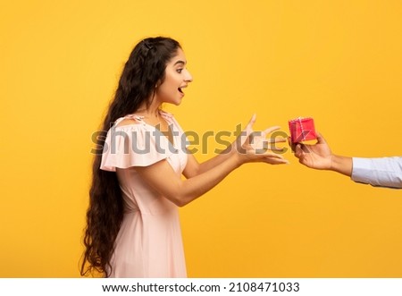 Holiday celebration concept. Male hand holding and giving wrapped gift box to surprised pretty indian woman standing over yellow studio background