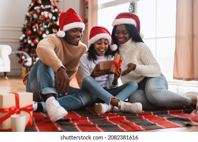 Holiday Celebration Concept. Portrait Of Happy Black Parents Giving Present To Excited Adolescent Daughter, Happy Girl Opens Gift Box, Family Greeting With Christmas And New Year Sitting On Floor