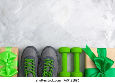 Holiday christmas birthday party sport flat lay composition with gray shoes, green dumbbells and craft gifts with green bow on gray concrete background. Top view, horizontal orientation