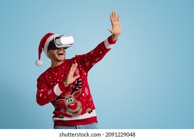 Holiday Entertainment. Young Excited Man In Santa Hat Using VR Headset, Joyful Man Wearing Christmas Sweater Experiencing Virtual Reality While Standing Over Blue Background In Studio, Copy Space