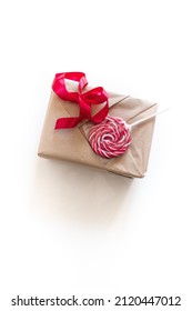 Holiday Gift Wrapping. Box Wrapped In Beige Kraft Paper With A Red Ribbon And A Red Striped Round Lollipop For Decoration. The Process Of Wrapping A Gift For Valentine's Day On A White Background