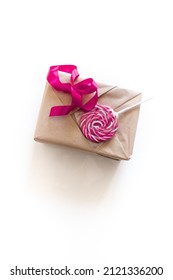 Holiday Gift Wrapping. Box Wrapped In Beige Kraft Paper With Pink Ribbon And Round Striped Pink Lollipop For Decoration. The Process Of Wrapping A Gift For Valentine's Day On A White Background. View