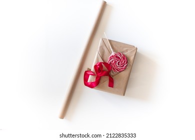 Holiday Gift Wrapping. The Box, Wrapped In Beige Kraft Paper, Lies Next To A Red Ribbon And A Round Lollipop For Decoration. The Process Of Wrapping A Gift For Valentine's Day On A White Background.