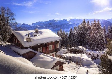 Holiday Home In The Winter With Snow Covering The Landscape In The Swiss Mountains With View On A Distant Mountain Range