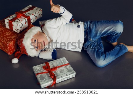 holiday photo with  little boy in Santa hat,jeans,white sweater smiling and lying with boxes and Christmas presents on the floor in fashion studio