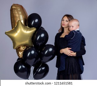 Holiday Photo Of Mom And Baby In Dark Clothes With Black And Gold Balls On A Light Background. The Concept Of A Busy Business Mom With A Baby In Her Arms