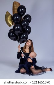 Holiday Photo Of Mom And Baby In Dark Clothes With Black And Gold Balls On A Light Background. The Concept Of A Busy Business Mom With A Baby In Her Arms