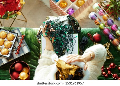 Holiday season. Upper view of unhappy trendy female in sparkle gold sequin santa hat sitting on divan in the modern living room at Christmas holding tangled christmas lights.