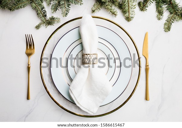 Holiday serving plate for Christmas eve. Beautiful\
white, light blue plates with golden rings, white napkin and golden\
fork and knife. Green Christmas tree branch. Concept of celebration\
table set.