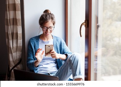 Holiday Shopping Online. Young Woman Makes Purchases In The Internet. Freelancer Pays By Using Smartphone On Black Friday. Happy Girl Is Enjoying Buying Gifts And Sitting By Window With Laptop At Home