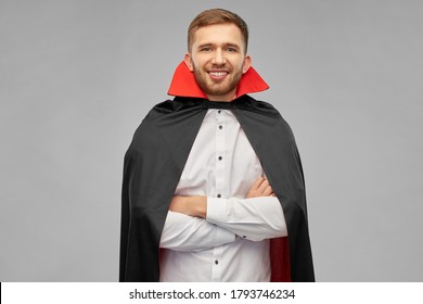 holiday, theme party and people concept - happy smiling man in halloween costume of vampire and dracula cape with arms crossed over grey background