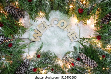 Holiday welcome sign with green Christmas tree garland border, pine cones, red berries, snow and lights on antique rustic wooden background; Christmas background with white copy space