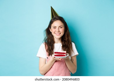 Holidays And Celebration. Cheerful Birthday Girl In Party Hat Holding Bday Cake And Smiling, Making Wish On Lit Candle, Standing Against Blue Background