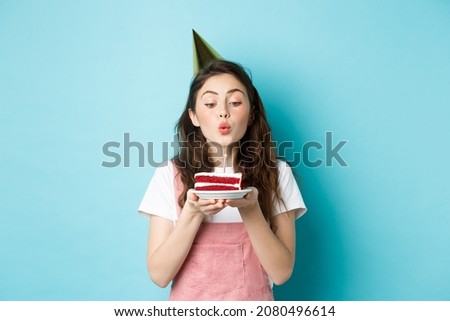 Holidays and celebration. Excited woman celebrating birthday, blowing candle on cake, wearing party cake and having fun, standing over blue background