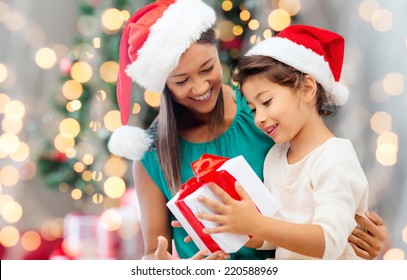 holidays, celebration, family and people concept - happy mother and little girl in santa helper hats with gift box over living room and christmas tree background