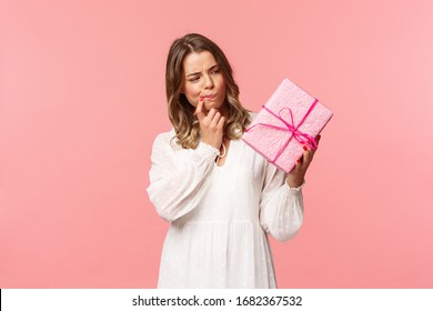 Holidays, Celebration And Women Concept. Portrait Of Thoughtful Curious Blond Girl Thinking Whats Inside Gift Box, Look Pondering Say Hmm As Making Assumpion, Standing Pink Background