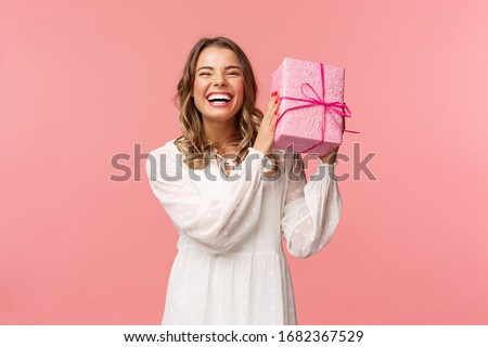 Holidays, celebration and women concept. Portrait of happy charismatic blond girl shaking gift box wondering whats inside as celebrating birthday, receive b-day presents, pink background