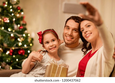 Holidays, Family And Technology Concept - Happy Mother, Father And Little Daughter With Christmas Gift Taking Selfie By Smartphone At Home